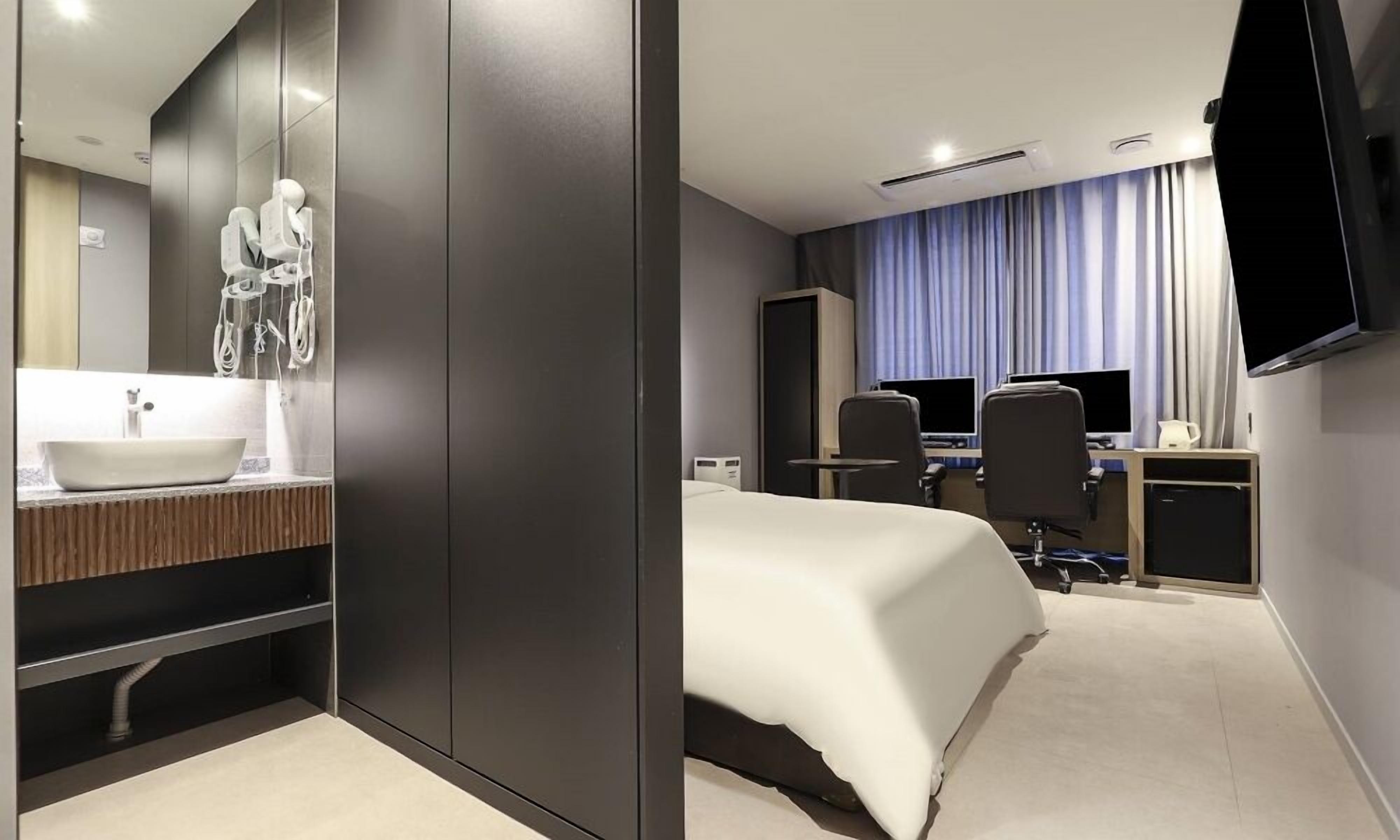 A modern hotel room with a large bed, a desk with two monitors, a flat-screen TV, and a bathroom with a sink and mirror.