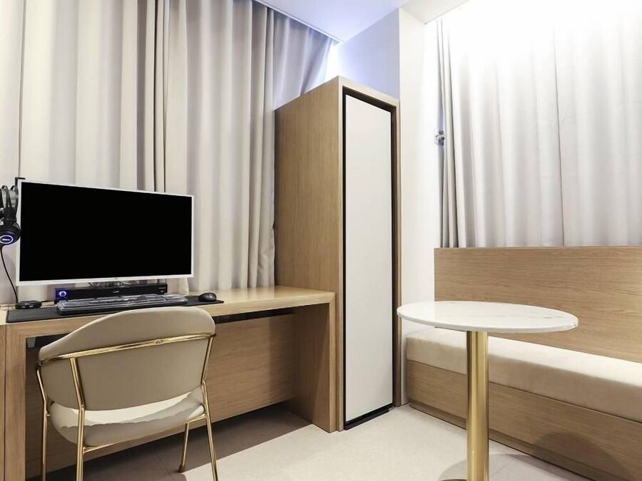 A modern hotel room with a wooden desk, a computer, headphones, a chair, a bed, and a small round table.