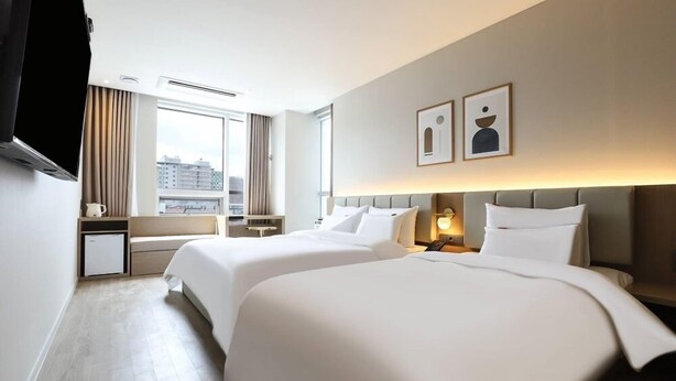 Une chambre d’hôtel avec deux lits, une télévision, une fenêtre avec des rideaux et des œuvres d’art accrochées au mur.
