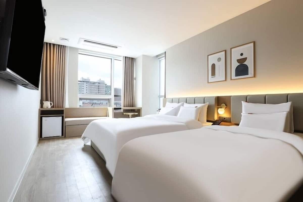 A hotel room with two beds, a TV, a window with curtains, and artwork on the wall.