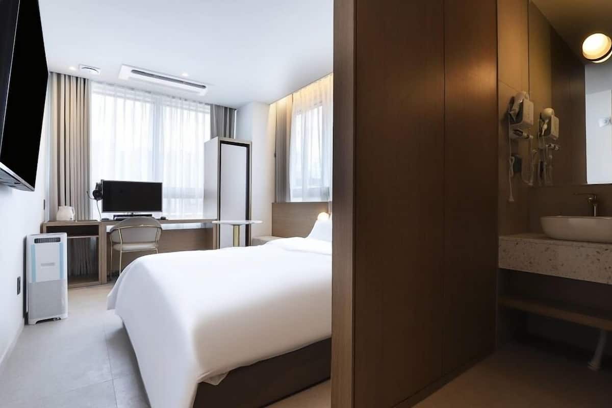 A hotel room with a bed, desk, chair, TV, and a bathroom with a sink and mirror.