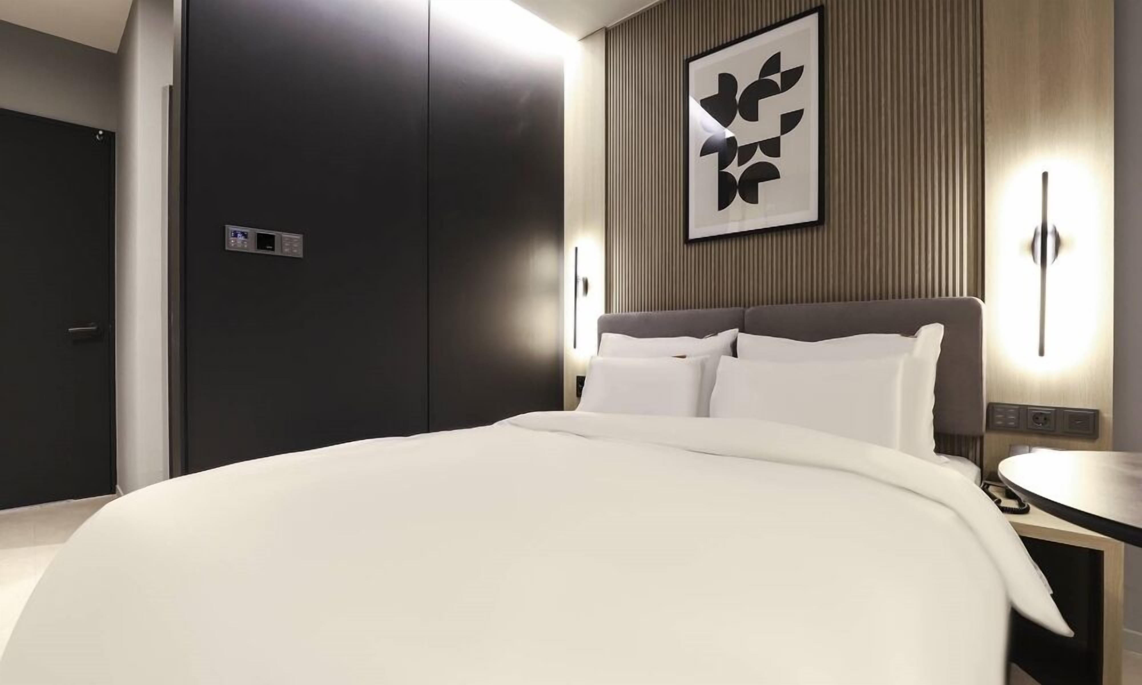 A modern hotel room with a large bed, a bedside table, a wall-mounted light, and a door with a control panel.