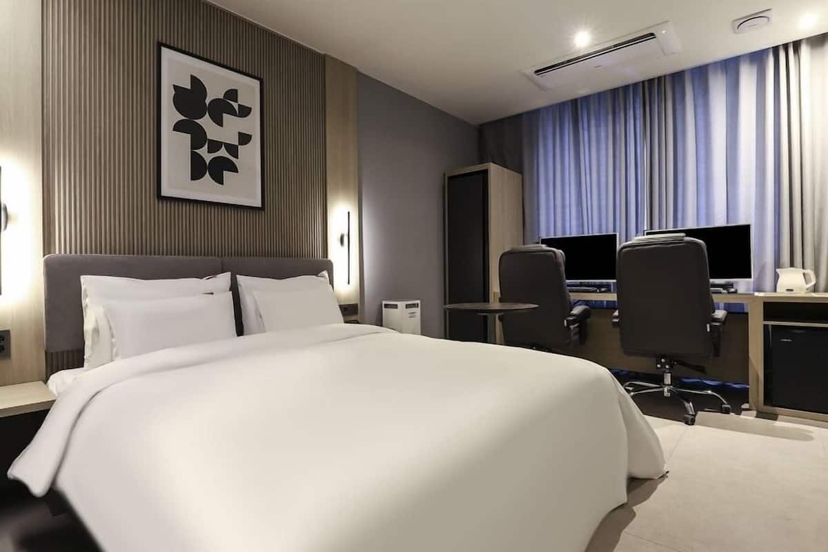 A modern hotel room with a large bed, a desk with two monitors, and a seating area with chairs.