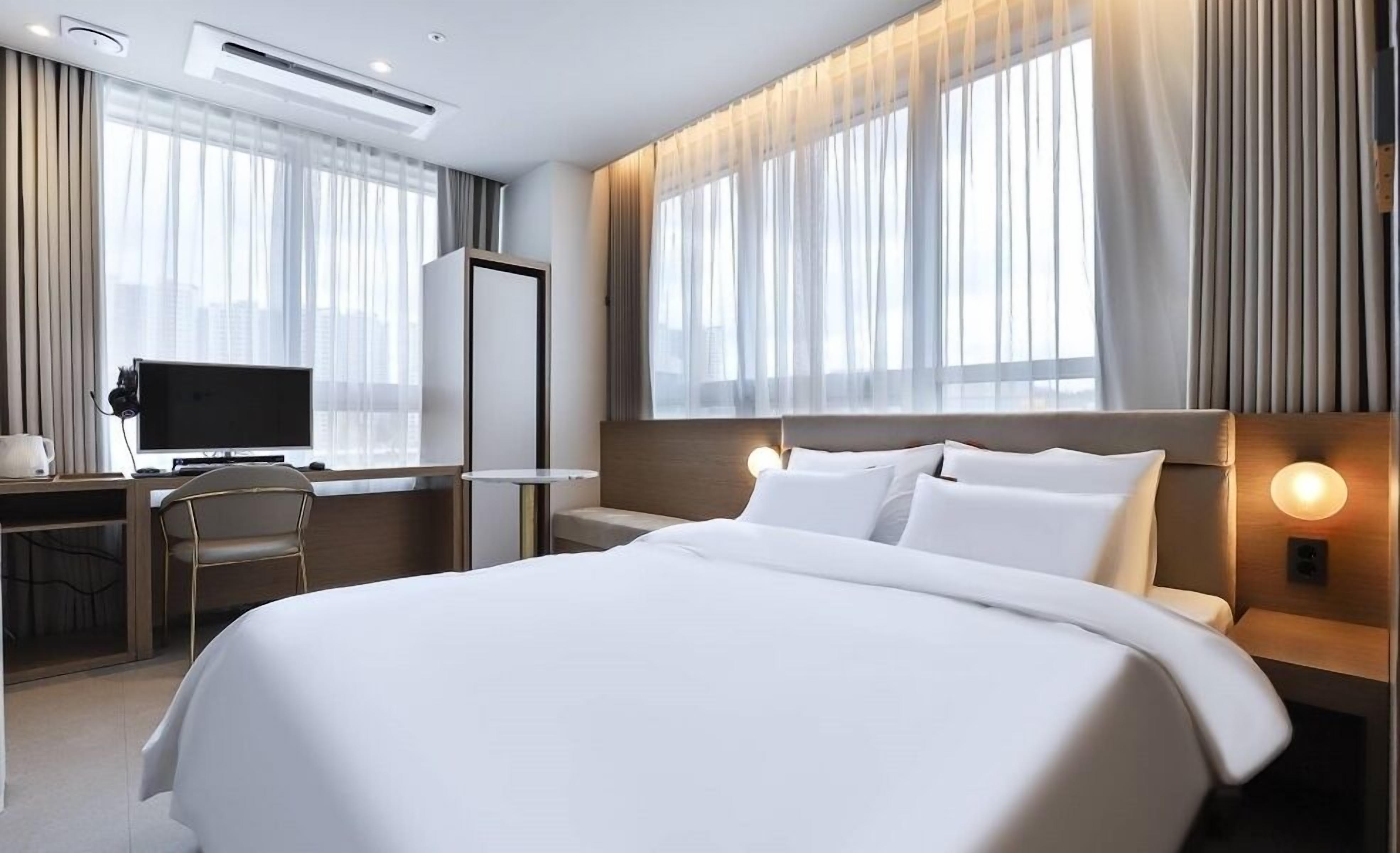 A hotel room with a large bed, a desk with a computer, and a chair. There are curtains on the windows, and a wall-mounted light fixture.