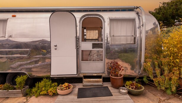 A stationary RV with a door open, revealing a small interior with a bed and a window. There are potted plants and a mat outside.