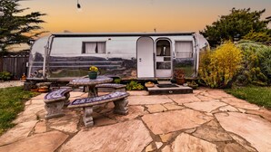 A stationary RV with a white exterior, a door, and a small window. It is parked on a stone patio with a table and benches.