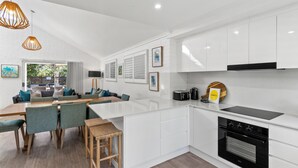 A modern kitchen with white cabinetry, a built-in oven, and a dining area with a wooden table and chairs. - Fuller Holidays - Somerset@Byron (Byron Bay)