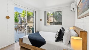 A bedroom with a wooden bed, a nightstand, a lamp, and a view of the outdoor patio. - Fuller Holidays - Somerset@Byron (Byron Bay)