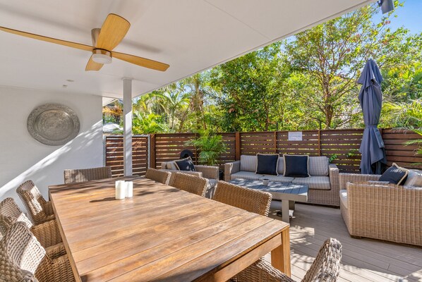 An outdoor patio with a wooden table and wicker furniture, surrounded by a wooden fence and lush greenery.