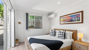 A bedroom with a bed, a nightstand, a lamp, a window with a view, and an air conditioning unit. - Fuller Holidays - Somerset@Byron (Byron Bay)