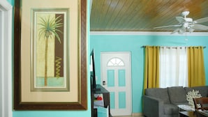 A living room with a teal wall, a framed picture of a palm tree, a grey sofa, a ceiling fan, and a door with yellow curtains.