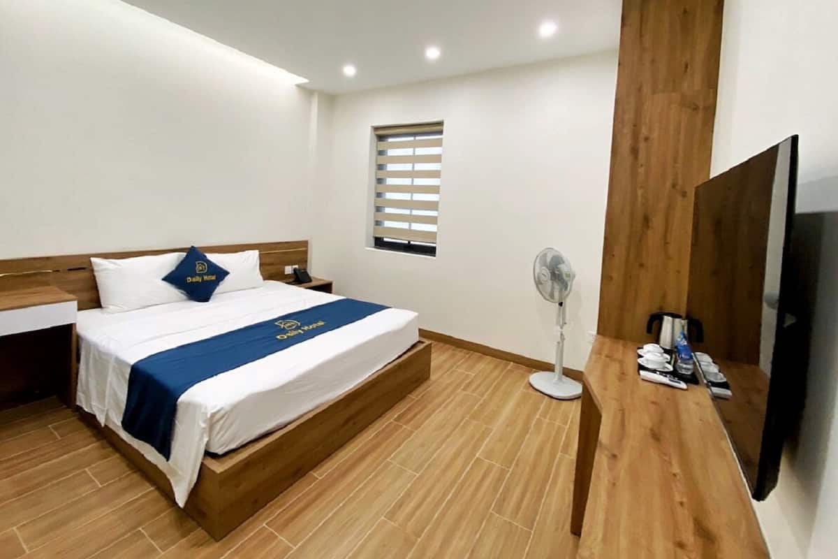 A hotel room with a bed, a bedside table, a TV, a fan, and a window with blinds.