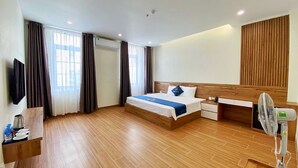 A modern hotel room with a wooden floor, a bed with a blue bedspread, a wooden headboard, a bedside table with a lamp, a TV mounted on the wall, and a standing fan. - Daily Hotel (Bai Chay)