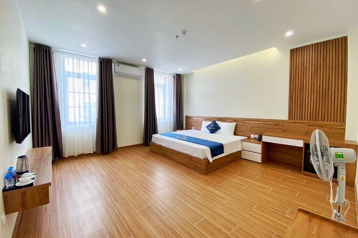 A modern hotel room with a wooden floor, a bed with a blue bedspread, a wooden headboard, a bedside table with a lamp, a TV mounted on the wall, and a standing fan.