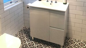 A bathroom with white tiled walls, a white sink with a mirror above it, a window, and a patterned floor. - Story Bank Mandurama   (Mandurama)