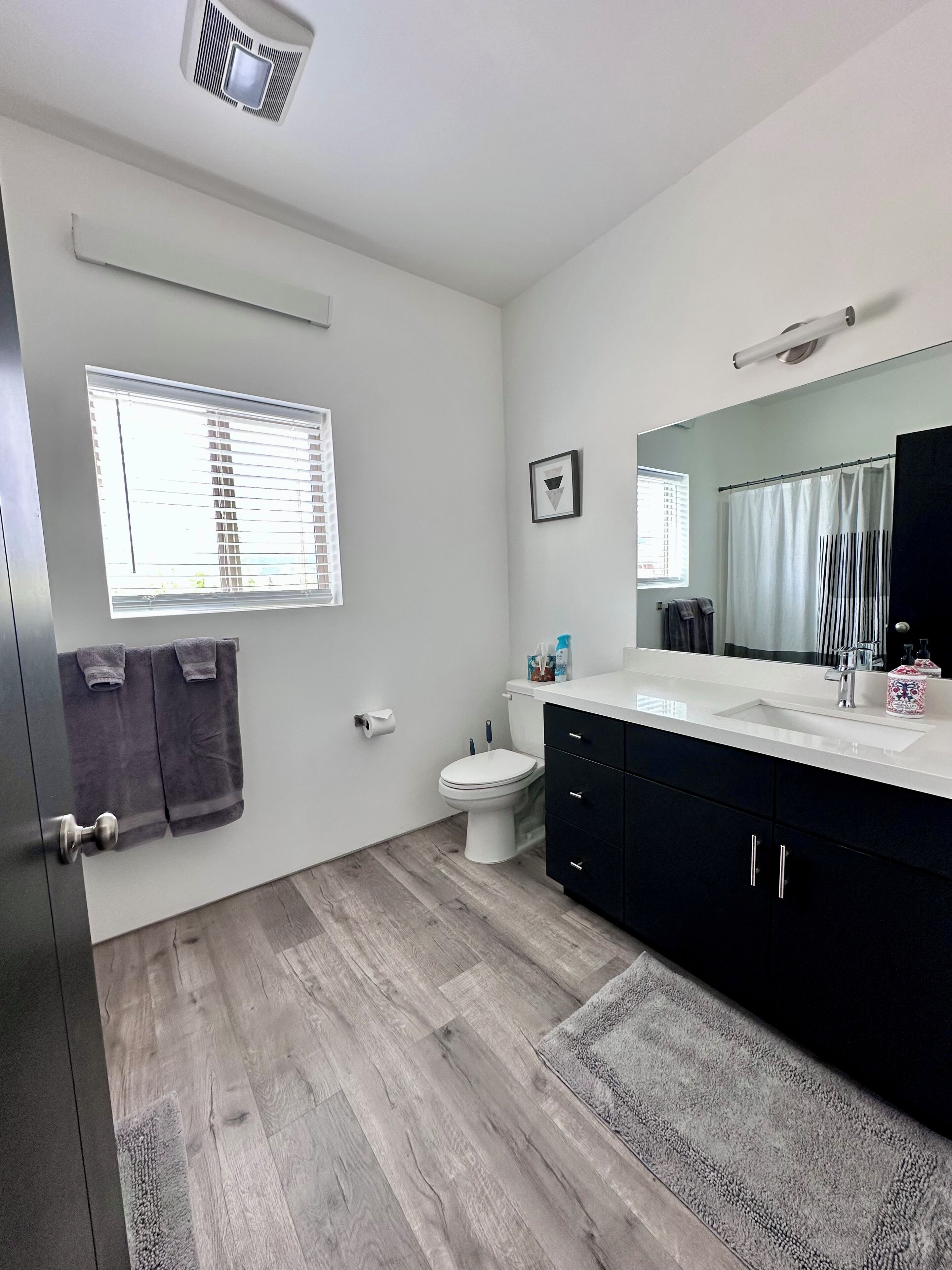 A modern bathroom with a toilet, sink, and dark cabinets. There is a window with blinds, a towel rack, and a picture on the wall.