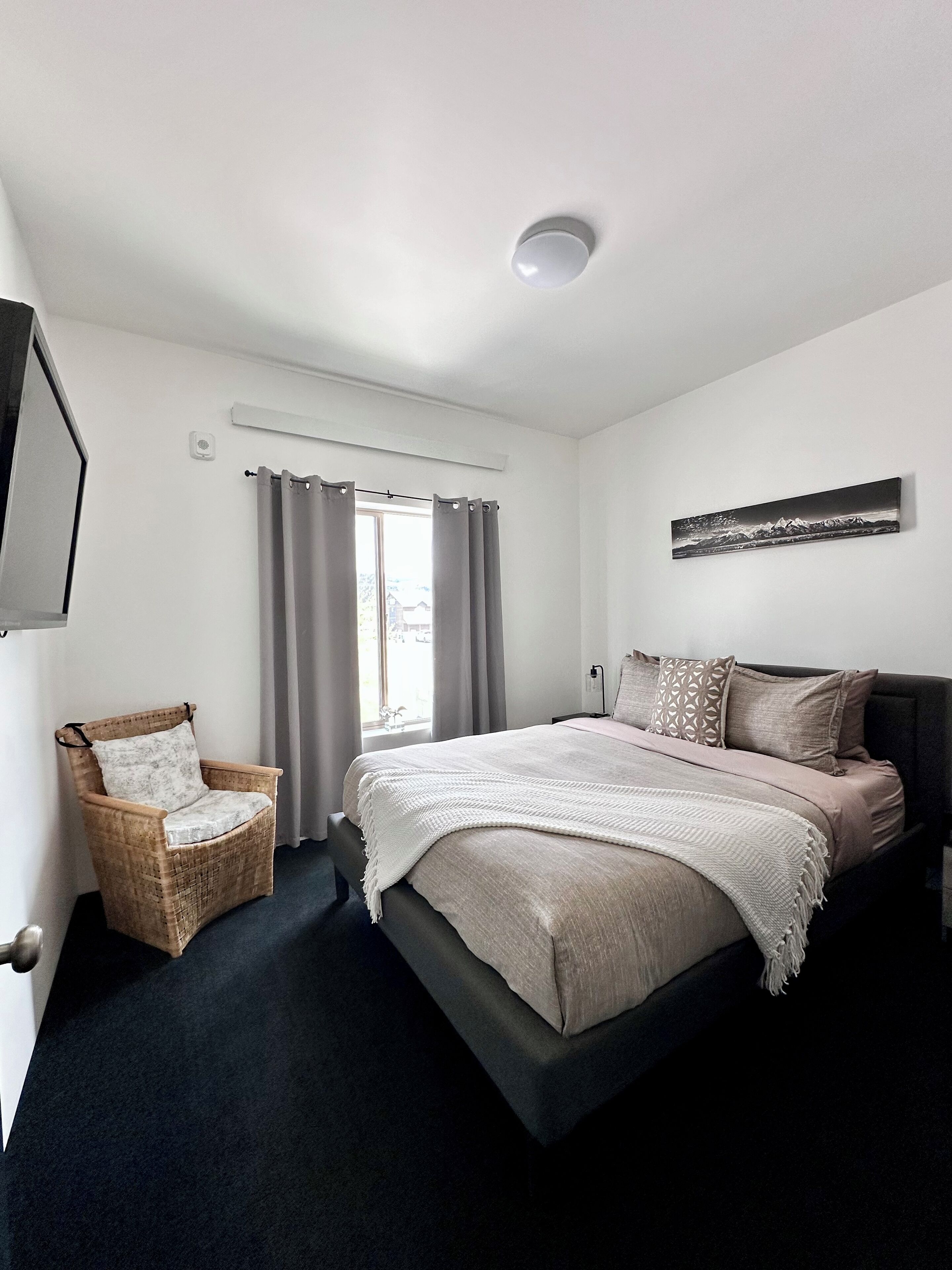 A bedroom with a bed, a chair, a window with curtains, a wall-mounted artwork, and a ceiling light.