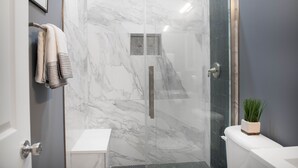 A modern bathroom with a glass shower enclosure, marble walls, a white sink, and a potted plant on the toilet. - Modern Mid-Town Luxury Hideaway  (Savannah)