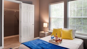 A hotel room with a bed, a nightstand, a lamp, a window with blinds, and a closet with hanging clothes. - Modern Mid-Town Luxury Hideaway  (Savannah)