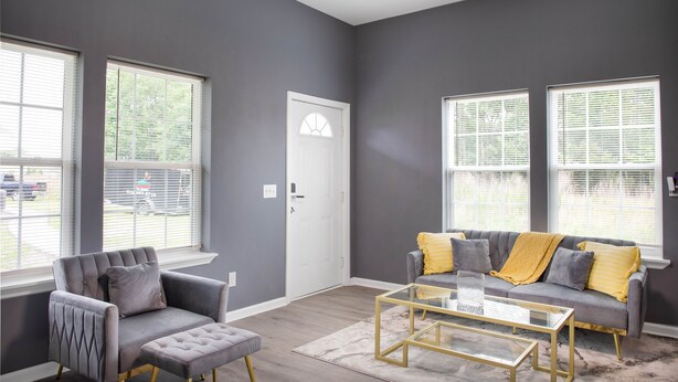 A modern living room with a grey sofa, armchair, and ottoman, a glass coffee table, and large windows with blinds. - Modern Mid-Town Luxury Hideaway  (Savannah)
