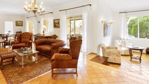 Un vaste salon comprenant un grand canapé, une table basse en verre et un lustre. On y trouve également un coin repas avec des chaises et une table, et une vue sur la verdure à l’extérieur.