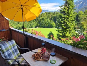 A balcony with a wooden table, a yellow umbrella, and a view of a green field and mountains. - Ferienwohnung Bergstüberl mit Südbalkon, 1-3 Personen, 50 qm (Bad Reichenhall)