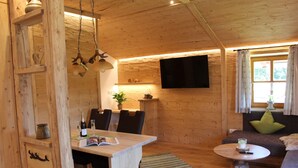 A wooden interior with a dining table, chairs, a sofa, a TV mounted on the wall, and a window with curtains. - Ferienwohnung Almstüberl mit Südbalkon, 1-3 Personen, 50 qm (Bad Reichenhall)