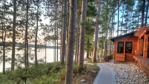 A cabin with a gravel path leading to a wooden deck, surrounded by tall pine trees and a lake view. - Newly Remodeled Lakefront Cabin w/ Modern Amenities. (Traverse City)