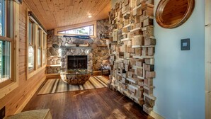 A cozy room with a stone fireplace, wooden walls, and a large window. - Newly Remodeled Lakefront Cabin w/ Modern Amenities. (Traverse City)