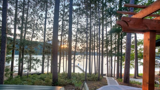 A wooden deck with a bench and a table overlooking a lake, surrounded by tall trees. - Newly Remodeled Lakefront Cabin w/ Modern Amenities. (Traverse City)
