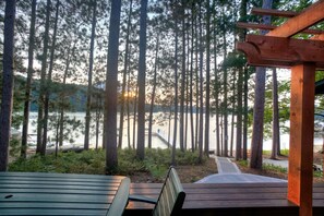 Una terraza de madera con un banco y una mesa, con vista a un lago rodeado de árboles altos.