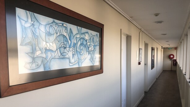 Een hal met een groot abstract schilderij aan de linkerwand, deuren aan de rechterkant, en een brandblusser aan de muur bevestigd.