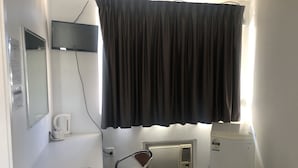 Een kleine hotelkamer met een enkel bed, nachtkastje met lamp, een klein verwarmingstoestel en een raam met een zwart gordijn.