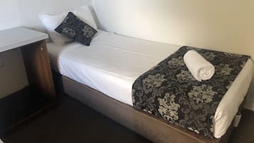 A single bed with a white mattress, a dark patterned bedspread, and a rolled white towel. There is a small wooden nightstand beside the bed.