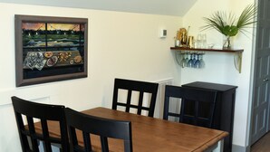 A dining area with a wooden table and black chairs, a painting on the wall, and a shelf with glasses and a plant.