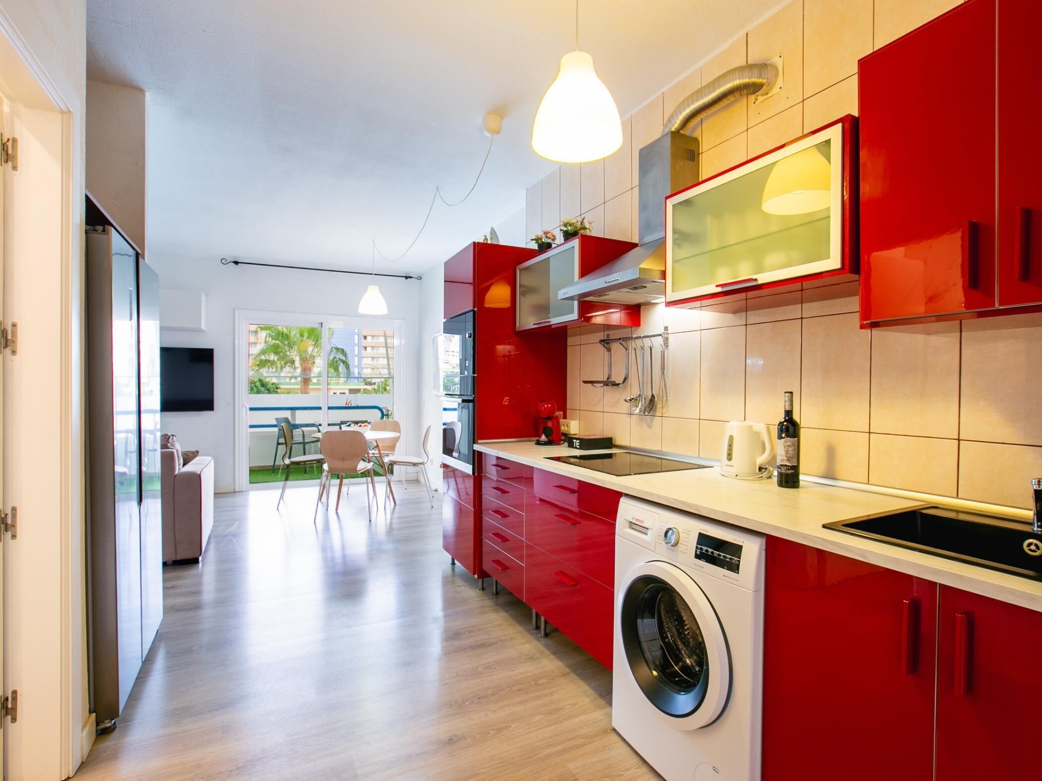 A modern kitchen with red cabinets, a built-in washing machine, and a dining area with a view.