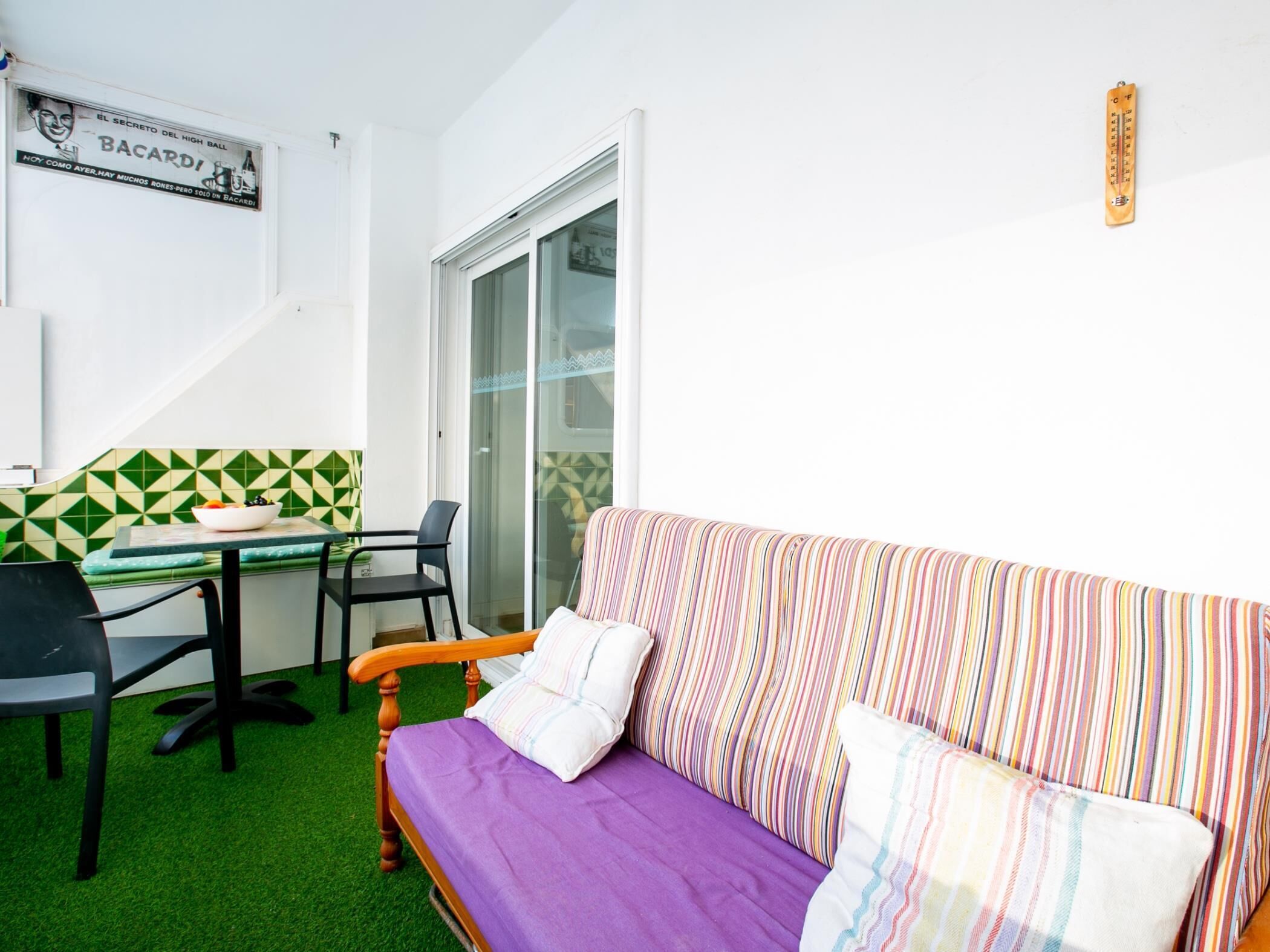 A small, brightly lit room with a purple sofa, a striped couch, a dining table with chairs, and a tiled backsplash.