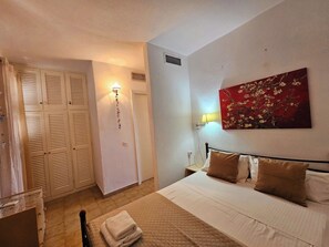 Une chambre à coucher avec un lit, des oreillers, une lampe fixée au mur, un tableau et une armoire à portes à persiennes.
