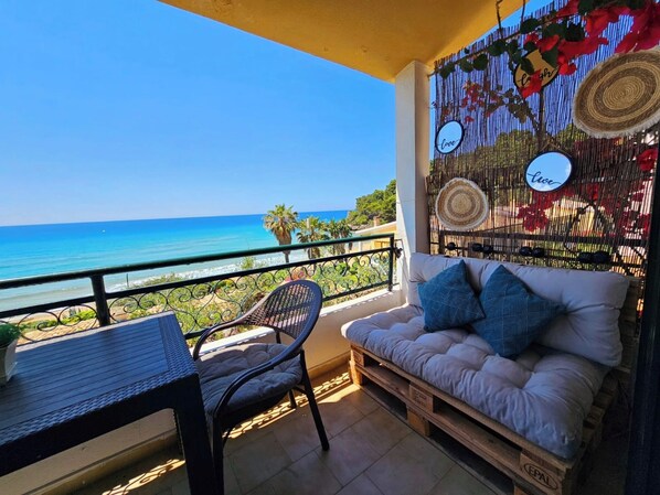 Un balcon avec vue sur la mer, une chaise en osier, un canapé avec des coussins et des éléments décoratifs suspendus au mur.