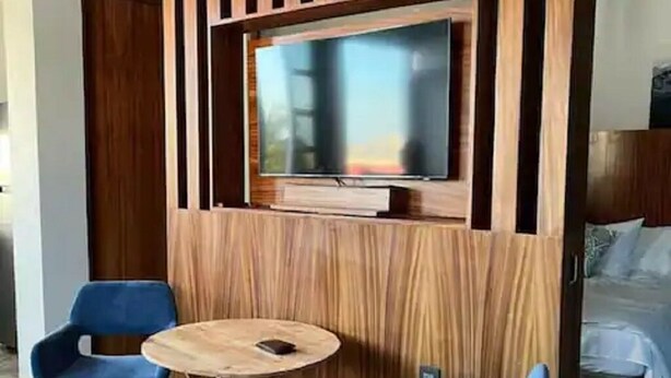 A room with a TV mounted on a wooden panel wall, two blue chairs, and a round wooden table.