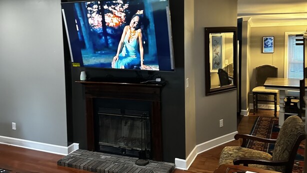 Een woonkamer met een flatscreen televisie boven een open haard, houten vloeren en een eethoek met een tafel en stoelen.