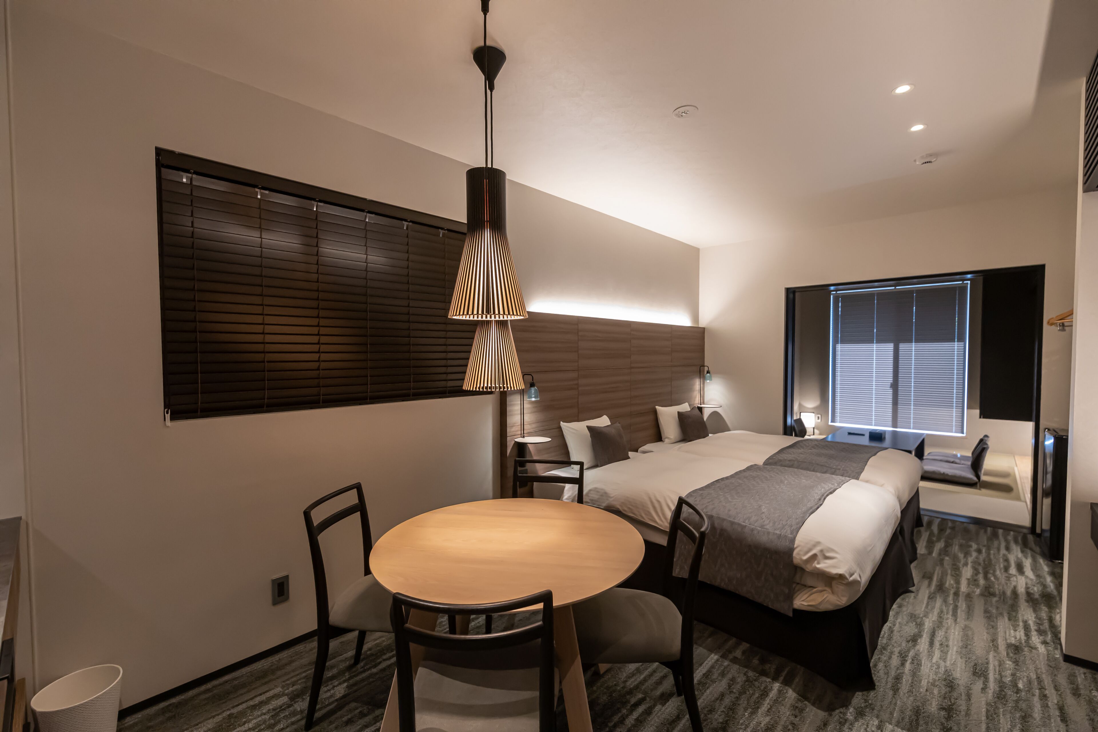 Ein modernes Hotelzimmer mit einem großen Bett, einem Esstisch mit Stühlen, einer kleinen Sitzecke und einem großen Fenster mit Jalousien.
