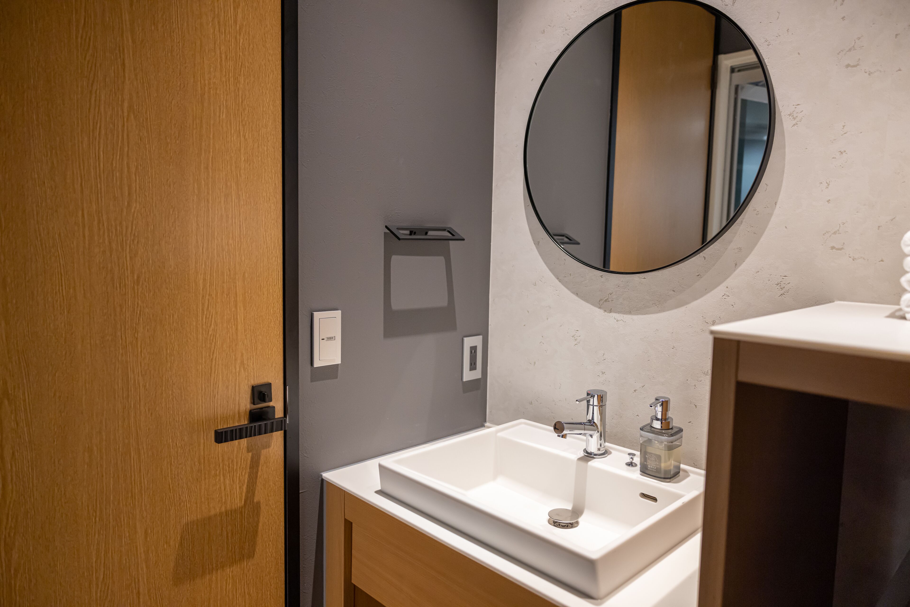 A modern bathroom with a wooden door, a white sink, a round mirror, and a small shelf.