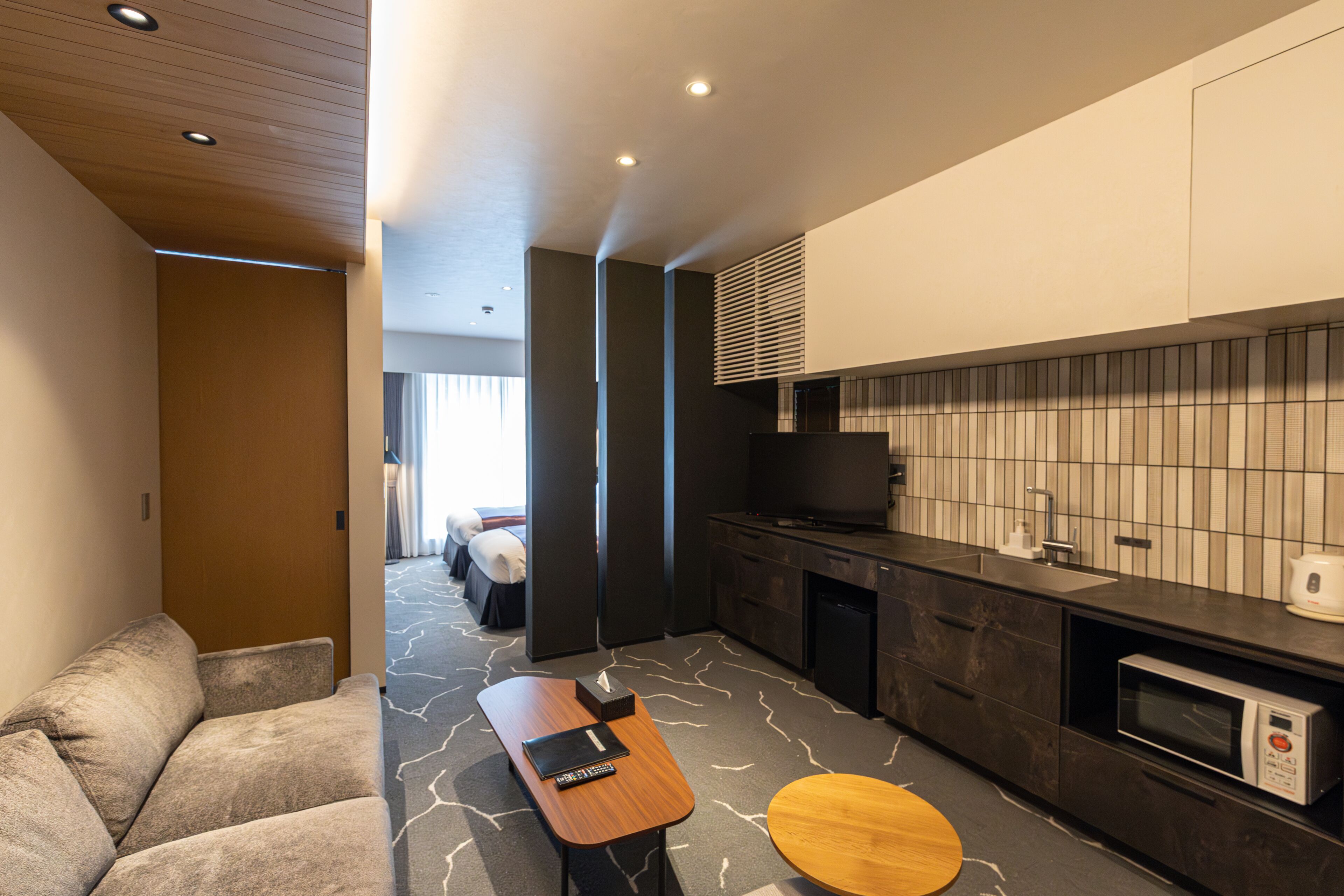 A modern hotel room with a sofa, a wooden side table, a small round table, a microwave, a TV, a sink, and a tiled backsplash.