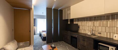 A modern hotel room with a sofa, a wooden side table, a small round table, a microwave, a TV, a sink, and a tiled backsplash.