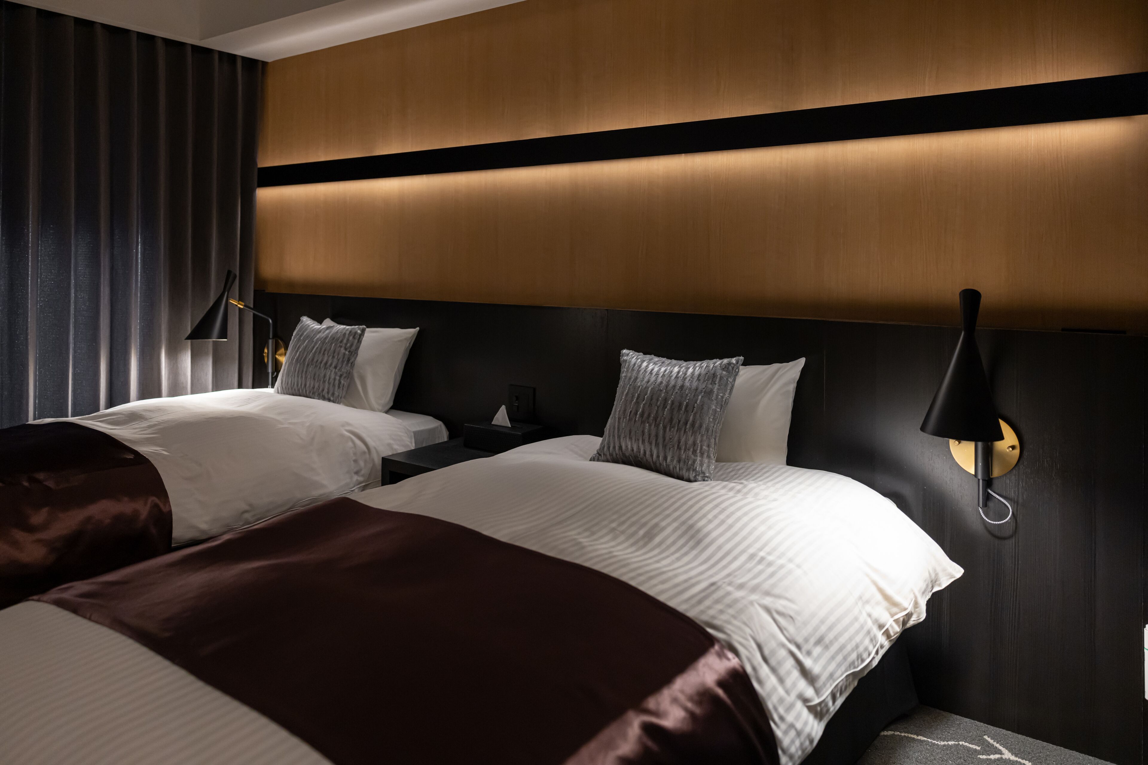 Habitación de hotel con dos camas, cabecera de madera oscura, iluminación moderna y una mesita de noche con lámpara.
