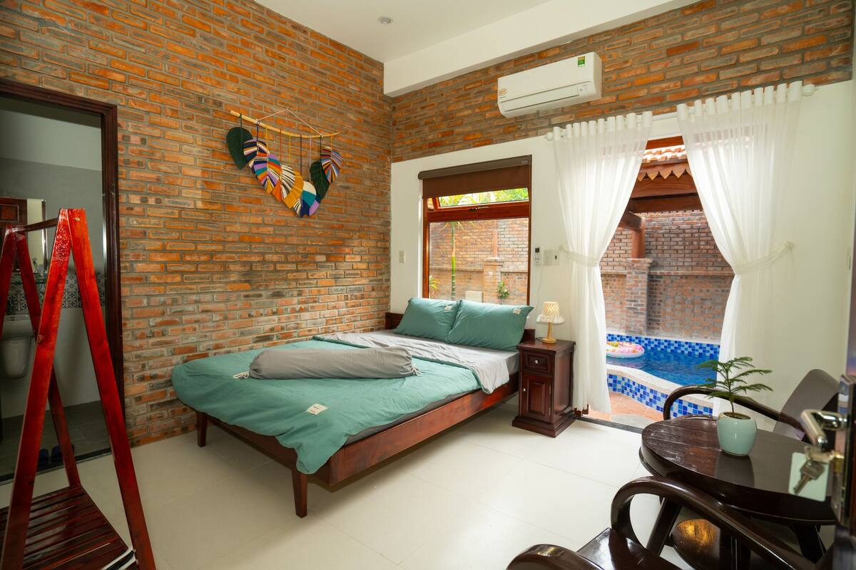 A bedroom with a brick wall, a bed with green bedding, a small table with a lamp, and a view of a swimming pool through a window.