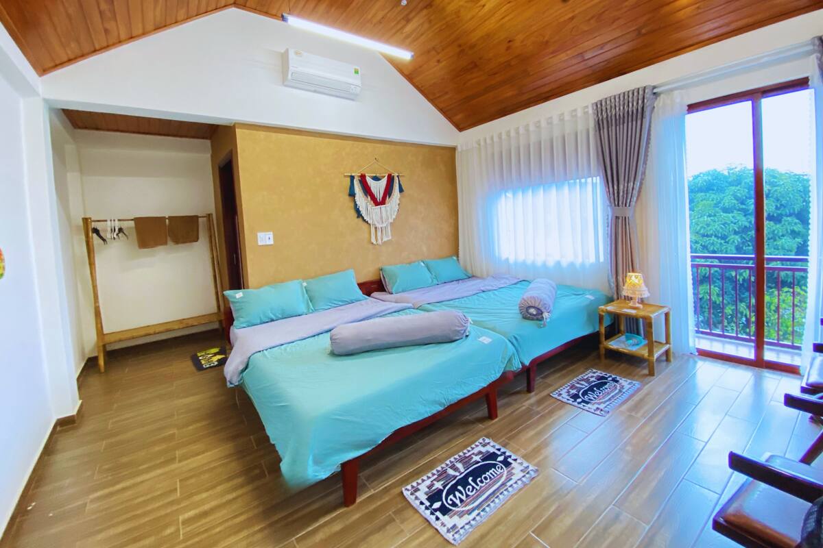 A bedroom with a wooden ceiling, a bed with teal bedding, a small bedside table with a lamp, and a balcony with a view of greenery.
