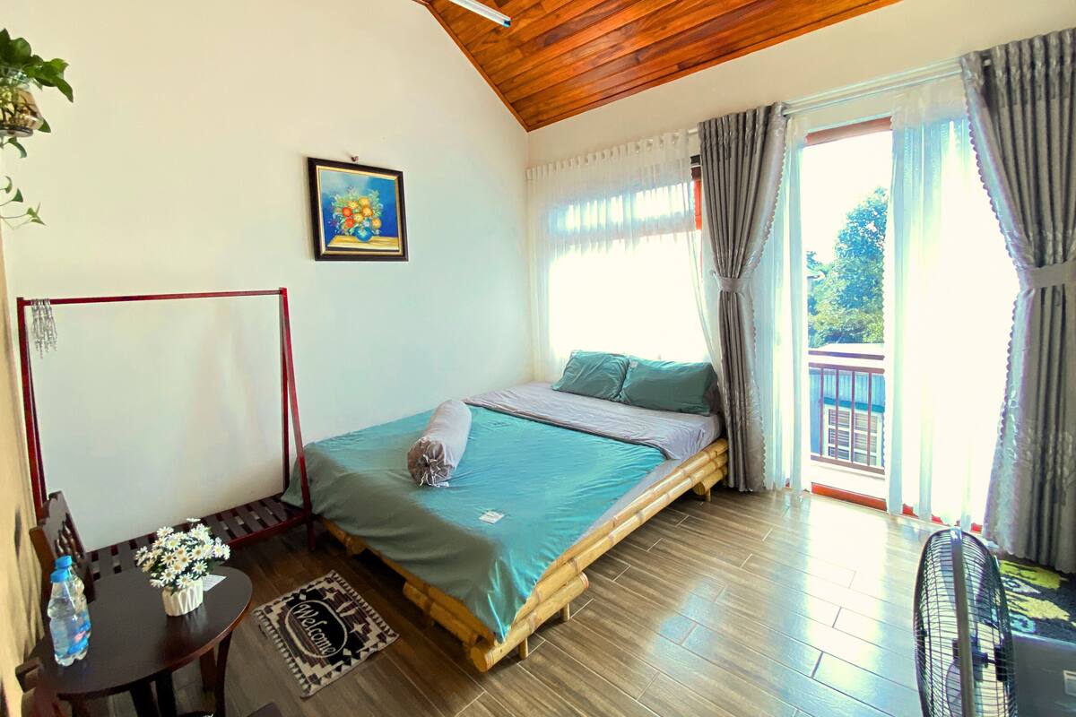 A room with a bed, a table with a bottle and flowers, a painting, and a view of the outside.