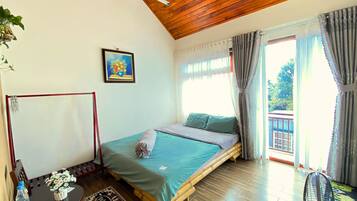 A room with a bed, a table with a bottle and flowers, a painting, and a view of the outside.